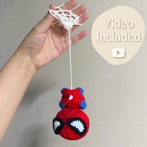 May include: A handmade crochet Spider-Man ornament, featuring a red body with black web detailing and white eyes. The ornament is suspended by a white string and a white web-like structure. The text "Video Included" is visible.