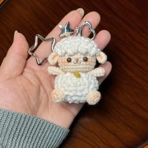 May include: A handmade white crochet sheep keychain with a silver star and heart-shaped keyring. The sheep has a gold star charm on its chest and light pink accents on its ears and feet. The keychain is held in a hand.