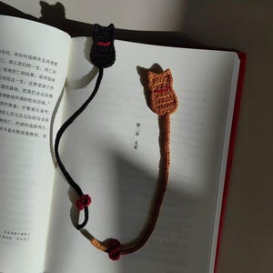 May include: Two handmade crochet cat bookmarks. One is black with a red collar, the other is orange with a red collar. The bookmarks are placed in an open book with red edges. The bookmarks have long tails with red accents.