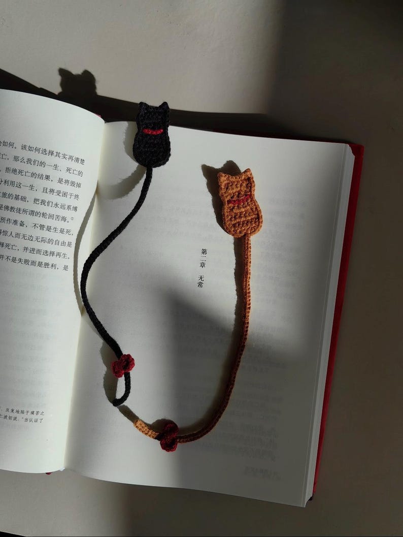 May include: Two handmade crochet cat bookmarks. One is black with a red collar, the other is orange with a red collar. The bookmarks are placed in an open book with red edges. The bookmarks have long tails with red accents.