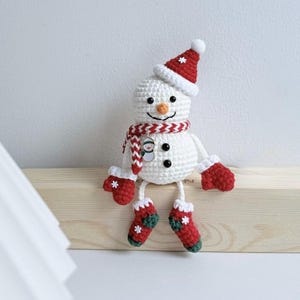 May include: A handmade crochet snowman wearing a red hat, scarf, mittens, and boots. The snowman has a carrot nose, black button eyes, and a smiling mouth. The scarf has a small snowman charm. A festive Christmas decoration.