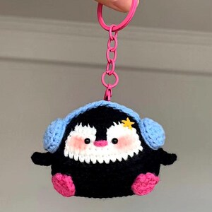 May include: A handmade crochet penguin keyring. The penguin is black with a white face, pink cheeks, and pink feet. It has blue earmuffs and a yellow star on its head. The keyring has a pink ring and chain.