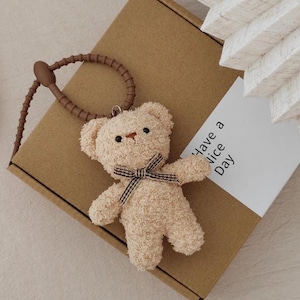 Op de afbeelding: Een kleine, beige teddybeer sleutelhanger met een bruine strik en een bruine riem. De beer heeft zwarte ogen en een bruine neus. Een witte kaart met de tekst "Have a Nice Day" is zichtbaar.