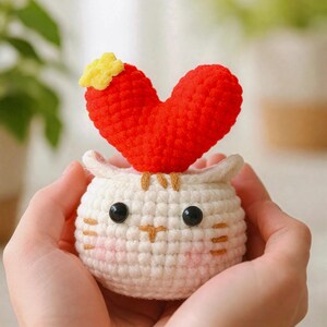 May include: A handmade crochet cat with a red heart on top. The cat is white with black eyes, pink cheeks, and brown whiskers. A small yellow flower is attached to the heart. The cat is a cute, whimsical decoration.