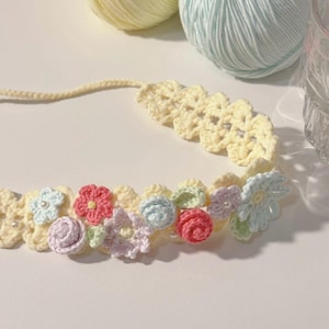 May include: A handmade crochet headband featuring a floral design. The headband is primarily cream-colored, with colorful crochet flowers in shades of pink, blue, and green. Small pearl beads are interspersed among the flowers. Two balls of yarn in pastel colors are in the background.