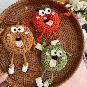 May include: Three amusing crochet food characters: a brown donut, a red tomato, and a green broccoli. Each has googly eyes, a pink tongue, and small white shoes. They are displayed on a woven tray, creating a playful and unique decorative item.