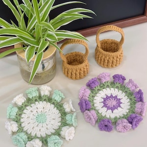 Puede incluir: Una colección de artículos de ganchillo hechos a mano, que incluyen dos pequeñas cestas y dos posavasos florales. Las cestas son de color marrón cálido, y los posavasos presentan diseños de flores blancas, verdes y moradas. Una planta en maceta está en el fondo.