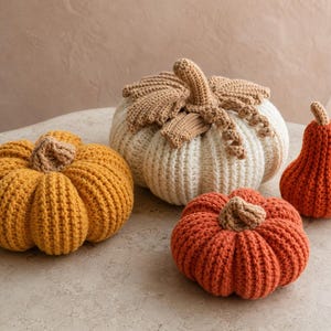 May include: A collection of four crocheted pumpkins in various sizes and colors. One large white pumpkin with tan leaves, one medium mustard yellow pumpkin, one medium orange pumpkin, and one small orange pear-shaped gourd, all with tan stems.