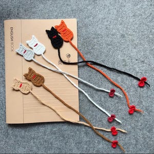 May include: A collection of crocheted cat bookmarks in shades of grey, white, black, orange, and brown. Each bookmark has a cat design with a red detail and a long cord, finished with a small red flower. The bookmarks are displayed on a book.