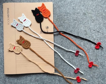 Könnte beinhalten: Gehäkelte Katzen-Lesezeichen in verschiedenen Farben wie Grau, Weiß, Schwarz, Orange und Braun. Jedes Lesezeichen zeigt eine Katzen-Silhouette mit roter Verzierung und einer langen Kordel, die in einer kleinen roten Blume endet. Auf einem Buch platziert.