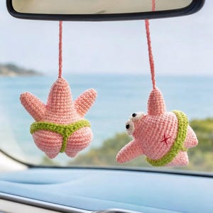 May include: Two crocheted pink starfish ornaments hanging from strings. Each figure features a bright green waistband, with one showing a rear view and the other showing a side profile with white eyes and a small red cross stitch on the side.