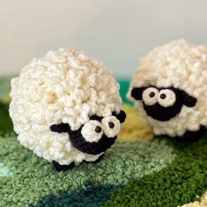 May include: Two handmade crochet sheep toys. The sheep are white with black faces, ears, and feet. They have large, googly eyes. The sheep are sitting on a green, yellow, and blue crocheted blanket.