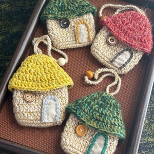 Puede incluir: Colección de cinco adornos de casa hechos a mano con ganchillo. Cada casa presenta un tejado de diferente color, incluyendo verde, rojo y amarillo. Las casas tienen detalles de botones y pequeños acentos de puerta. Se exhiben en una bandeja marrón.