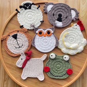 May include: A collection of seven handmade crochet coasters in various animal shapes, including a sheep, koala, lion, owl, chicken, dog, and frog. Each coaster features detailed stitching and vibrant colors, set on a wooden tray.