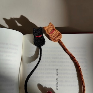May include: Two handmade crochet cat bookmarks. One is black with a red collar, the other is tan with a red mouth. The bookmarks are lying on an open book. The bookmarks are connected to a decorative red and brown floral accent.
