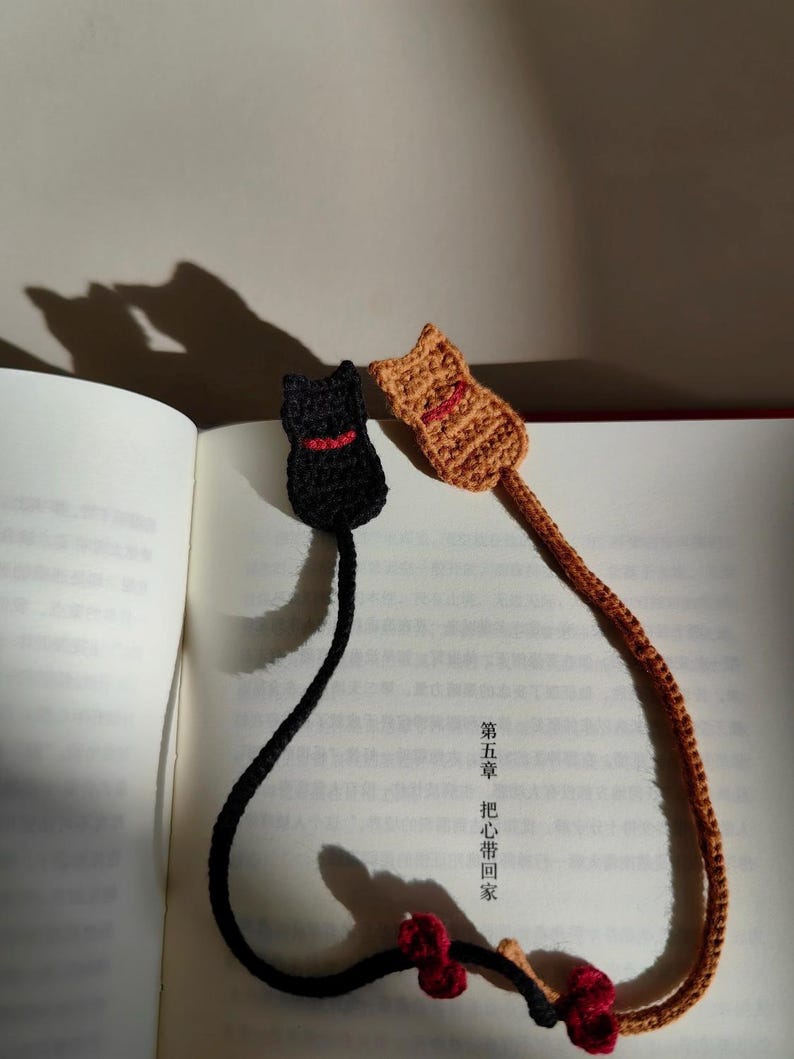 May include: Two handmade crochet cat bookmarks. One is black with a red collar, the other is tan with a red mouth. The bookmarks are lying on an open book. The bookmarks are connected to a decorative red and brown floral accent.