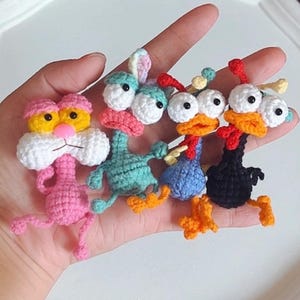 May include: A selection of handmade crochet animal figures, including a pink cat, a teal duck, and two chickens. The figures have large, expressive eyes and are made with colourful yarn. The cat has yellow eyes and a white muzzle. The chickens are black and red.