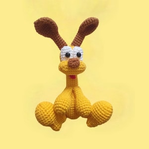 May include: A handmade crochet dog toy in yellow, brown, white, and black. The dog has large brown ears, white and black eyes, and a red collar. The toy is sitting on a yellow background.