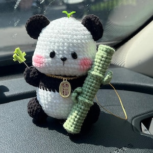 May include: Crocheted panda bear car charm with black and white body, black ears, and pink cheeks. The panda holds a green crocheted bamboo stalk and a small green sprig. A gold-colored charm with Chinese characters hangs from its neck.