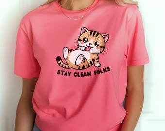 Può includere: Una maglietta rosa con un gatto dei cartoni animati arancione e marrone, sdraiato sulla schiena con la lingua di fuori. Il testo "STAY CLEAN FOLKS" è stampato sotto. La maglietta ha maniche corte e scollo tondo.