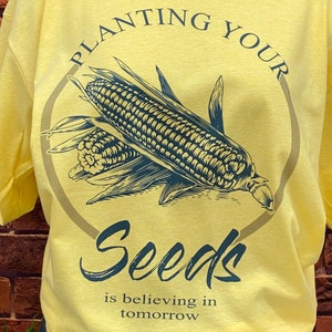 May include: Yellow t-shirt with a black and white illustration of corn and the text "Planting Your Seeds is believing in tomorrow".