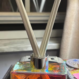 May include: A square glass diffuser bottle with a holographic finish, filled with orange liquid and white reeds. The bottle has a silver collar and a label that reads "Pineapple Red Diffuser".