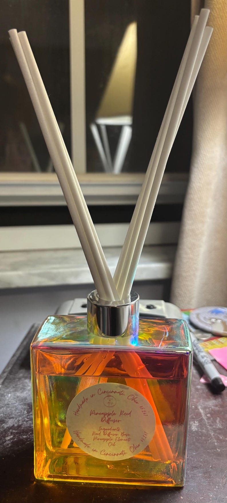 May include: A square glass diffuser bottle with a holographic finish, filled with orange liquid and white reeds. The bottle has a silver collar and a label that reads "Pineapple Red Diffuser".