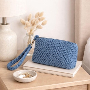 Può includere: Una pochette rettangolare in uncinetto blu con un motivo a punti testurizzati e un cinturino da polso blu abbinato fissato a una cerniera in metallo. La pochette poggia su un libro bianco, accompagnata da un piattino in ceramica con anelli.