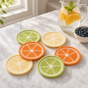 May include: A set of six circular crochet coasters designed to resemble citrus fruit slices. The collection includes two lime green, two bright orange, and two pale yellow coasters, each featuring a white center and white radial segment lines.