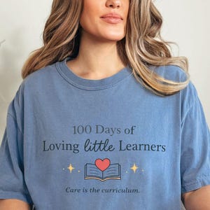 May include: A blue jean-colored t-shirt with the text "100 Days of Loving little Learners" and "Care is the curriculum." The design includes an open book with a red heart and star accents. The shirt is worn with ripped jeans.