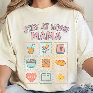 May include: Ivory-colored t-shirt with the text "STAY AT HOME MAMA" in pink and light blue. The shirt features nine square illustrations of baby-related items, including a pacifier, teddy bear, and bread.