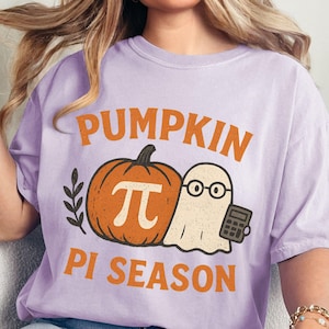 May include: A light purple t-shirt with the words "PUMPKIN PI SEASON" in orange. The graphic includes a pumpkin with a pi symbol, a ghost wearing glasses, and a calculator. The shirt is a comfortable fit.