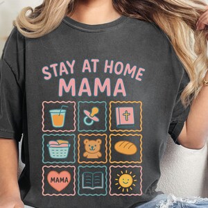 May include: Dark gray t-shirt with the text "STAY AT HOME MAMA" and colorful illustrations of a drink, pacifier, cross, basket, teddy bear, bread, heart, book, and sun. The shirt is a comfortable, casual style.