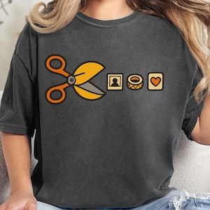 May include: Dark gray t-shirt featuring a graphic design with yellow scissors, a framed person, a spool of thread, and a heart. The shirt is a comfortable, casual style, perfect for everyday wear. The word "Pepper" is in the upper right corner.