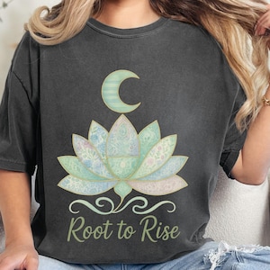 May include: Dark gray t-shirt featuring a lotus flower design with a crescent moon above it. The lotus is in shades of green and blue, with the words "Root to Rise" in a script font below the flower. The shirt is worn by a person.