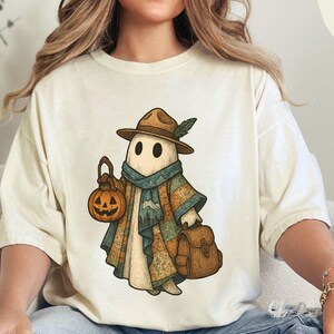 May include: Ivory-colored t-shirt featuring a whimsical ghost character dressed in a hat, scarf, and coat, carrying a pumpkin lantern and a satchel. The ghost has two black eyes and a friendly expression. The shirt is a relaxed fit.