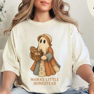 May include: Ivory-colored t-shirt featuring a whimsical illustration of a ghost character holding a basket of bread and a bottle. The ghost is dressed in a patchwork-style outfit with brown, orange, and beige tones. The text "MAMA'S LITTLE HOMESTEAD" is printed below the graphic.