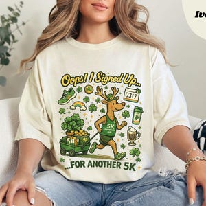 May include: Ivory-colored t-shirt with a graphic design featuring a running deer wearing a green running vest, a green running shoe, a rainbow, and shamrocks. The text on the shirt says "Oops! I Signed Up... For Another 5K."