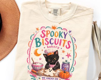 Op de afbeelding: Een crèmekleurig T-shirt met een afbeelding van een zwarte kat met een koksmuts die deeg in de vorm van een spookje kneedt. De tekst luidt "SPOOKY BISCUITS bakery" en "WE KNEAD EM YOU NEED EM", met illustraties van een meelpot en een pompoen.