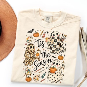 May include: A cream-colored t-shirt with a graphic print featuring three ghosts, pumpkins, paint palettes, brushes, and the text 'Tis the Season' in black script. The ghosts have leopard print, checkered, and star patterns.