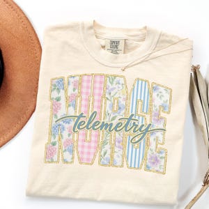May include: Cream-colored t-shirt with the words "NURSE TELEMETRY" in a collegiate-style design. The letters are filled with floral patterns, pink gingham, and blue stripes. A gold necklace and a brown hat are visible.