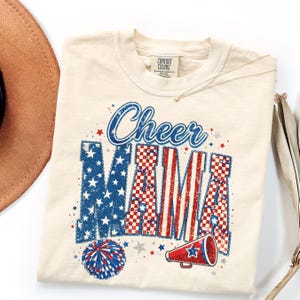 May include: Cream-colored t-shirt with the words "Cheer MAMA" in blue, red, and white. The design incorporates stars, a pom-pom, and a megaphone, all in patriotic colors. The shirt has a crew neck and a small tag.