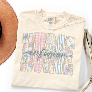 May include: Cream-colored t-shirt with the words "NURSE infusion NURSE" in a collegiate style. The letters are decorated with floral, gingham, and striped patterns. A gold necklace and a brown hat are visible.