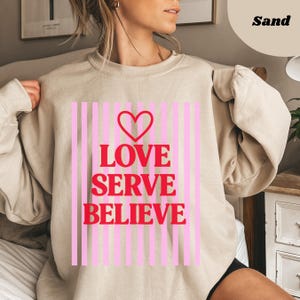 May include: A sand-colored sweatshirt with a pink and white striped design. The sweatshirt features the words "LOVE SERVE BELIEVE" in red, with a red heart above the word "LOVE".