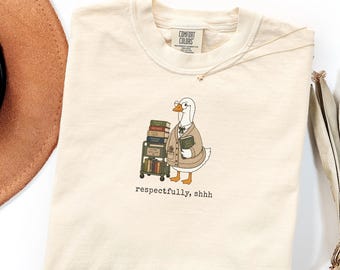 May include: Cream-colored Comfort Colors t-shirt featuring a cartoon goose wearing glasses and a blazer, pushing a cart of books. The text "respectfully, shhh" is printed below the goose. A brown hat and necklace are visible.