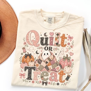 May include: A cream-colored t-shirt featuring a graphic with the text Quilt OR Treat. The design includes a ghost wrapped in a patchwork quilt, pumpkins, spools of thread, needles, bows, and hearts in shades of pink, orange, and brown.