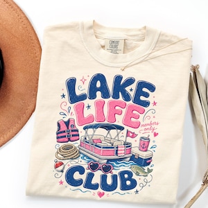 May include: A cream-colored t-shirt featuring a graphic print with the text "LAKE LIFE CLUB" in blue and pink. The design includes a pontoon boat, a life vest, a rope, a cooler, a drink, heart-shaped sunglasses, and a fish in blue and pink.