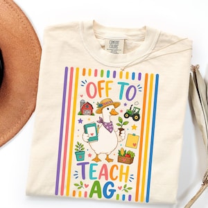 May include: A cream-colored t-shirt featuring a graphic of a white goose wearing a straw hat and purple bandana. The goose holds a notebook and a small plant. Surrounding the goose are a red barn, a green tractor, and the text OFF TO TEACH AG.