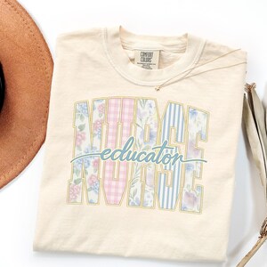 May include: Cream-colored t-shirt with the words "NURSE EDUCATOR" in a collegiate style. The letters are filled with floral and striped patterns in pink, blue, and white. A small gold necklace is visible.
