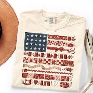 May include: A cream-colored t-shirt featuring a graphic design of an American flag composed of sewing-themed elements. The design includes red and blue quilt patterns, spools of thread, scissors, a measuring tape, and a sewing machine.